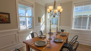 Comedor con mesa de madera dispuesta para cuatro personas, rodeado de sillas negras. Hay dos ventanas con persianas, una lámpara de candelabro y un cuadro en la pared.