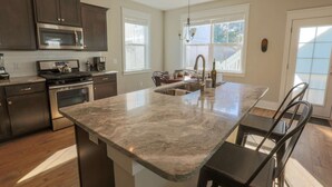 Una cocina con armarios oscuros, una estufa de acero inoxidable, una isla grande con un fregadero, y una zona de comedor con mesa y sillas.