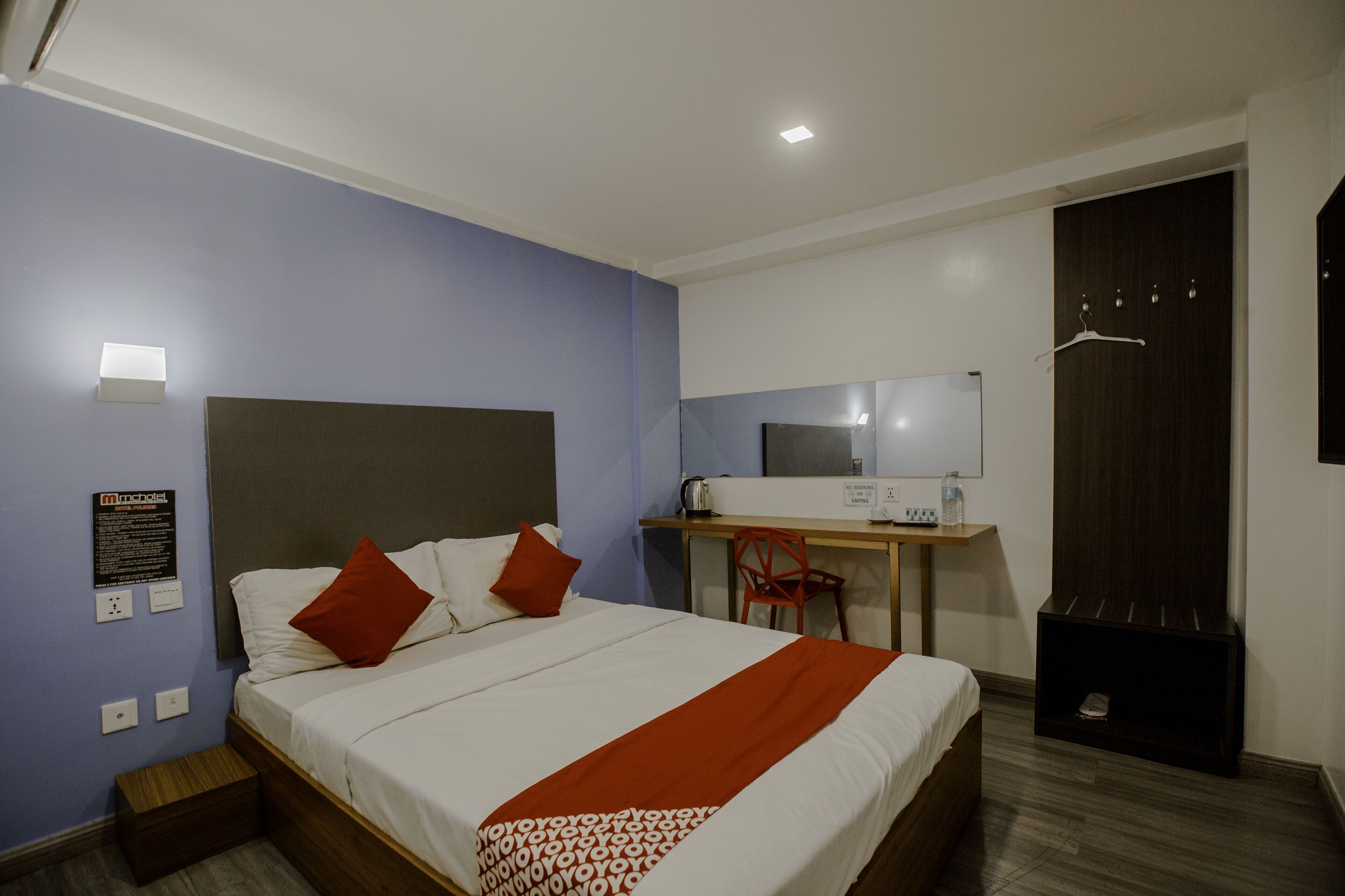 A hotel room with a bed, a desk, a chair, a mirror, a wardrobe, and a small wooden box.