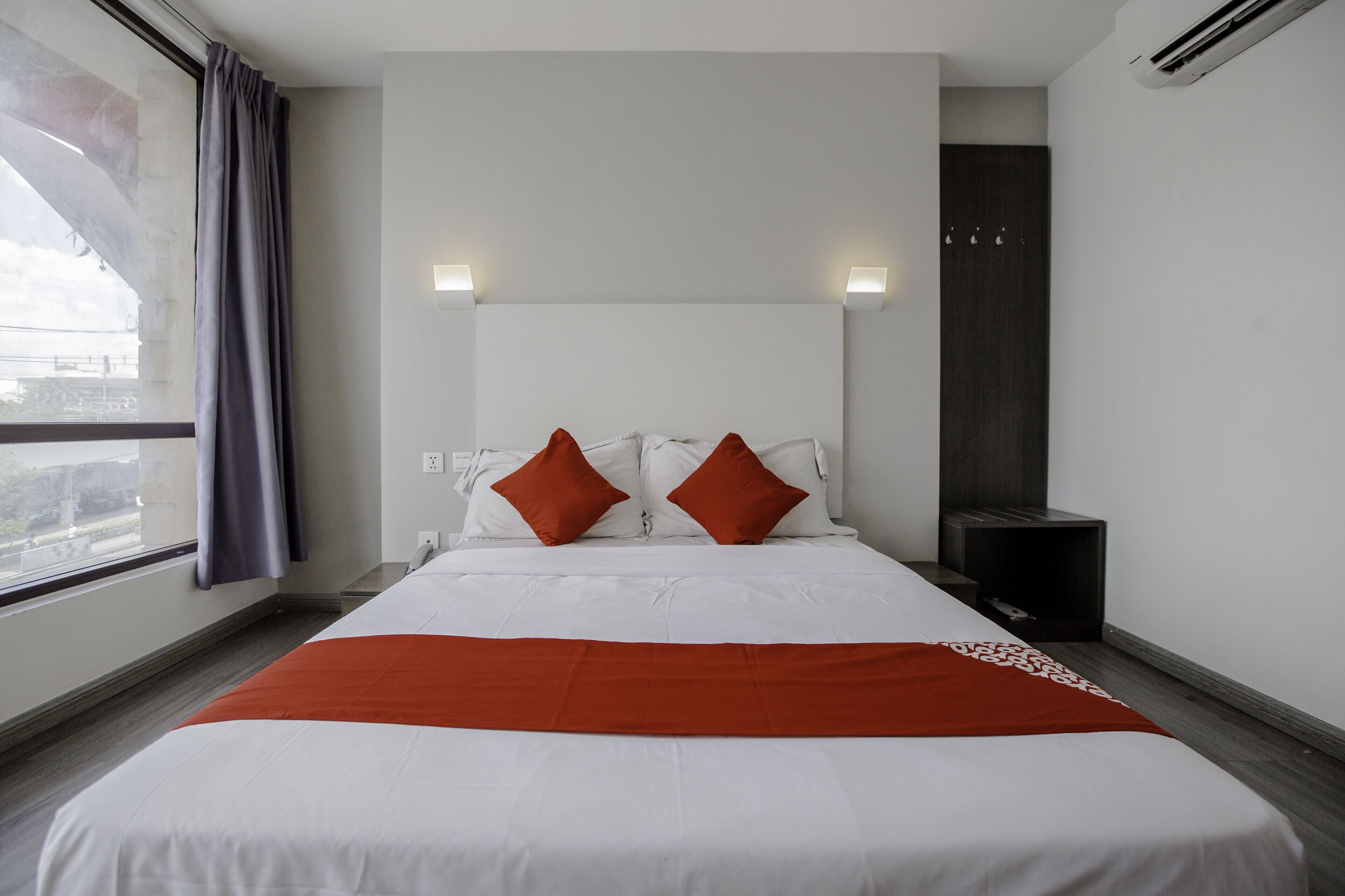 A modern hotel room with a large bed, two pillows, a red throw, and a view of the city through a window.