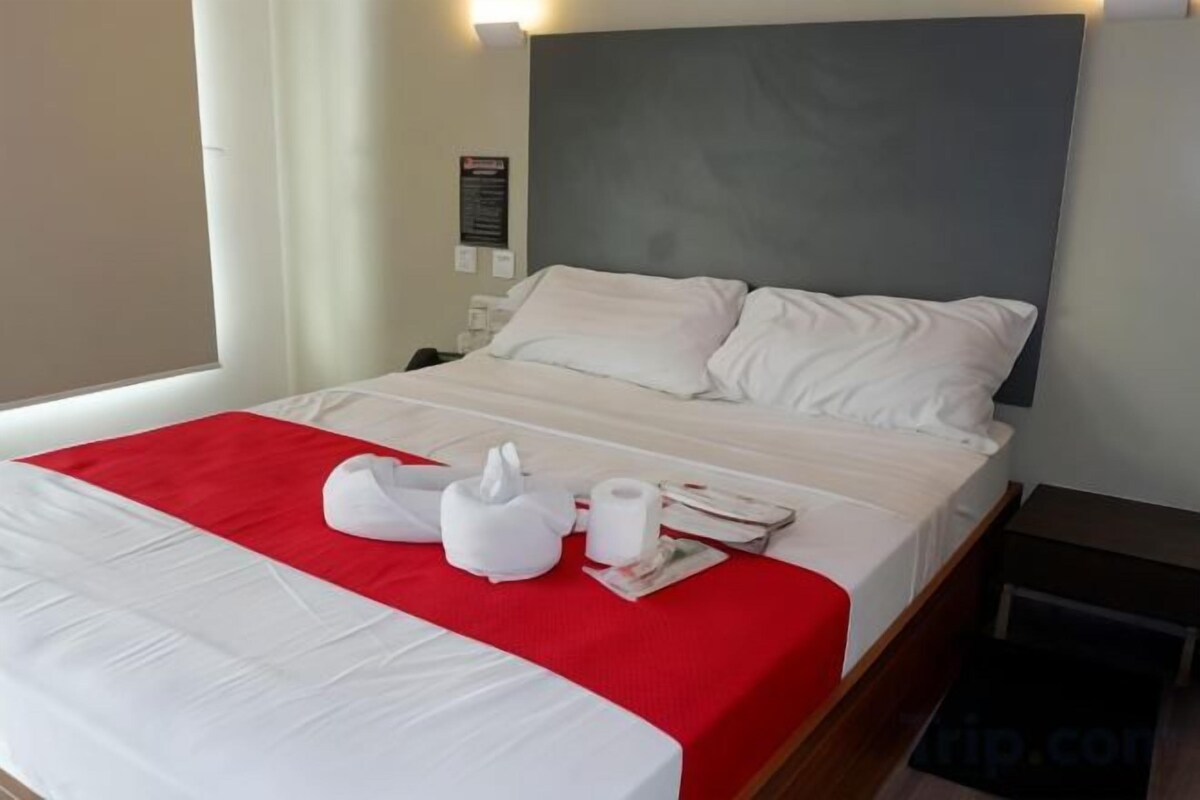 Hotelkamer met een bed, witte lakens, een rode plaid, een kussen, een zakdoekendoos en een afstandsbediening.