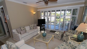 A living room with a sofa, coffee table, and patterned ottomans. A television is on a stand, and there is a ceiling fan. - Bayside 2BR on Whitney Beach w Canal Views, Pool, and Private Lanai  by RVA (Longboat Key)
