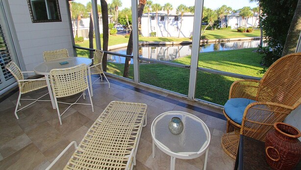 A sunlit patio with wicker furniture, a ceiling fan, and a view of a grassy area and water feature. - Bayside 2BR on Whitney Beach w Canal Views, Pool, and Private Lanai  by RVA (Longboat Key)