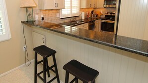 A kitchen with white cabinets, a granite countertop, a microwave, an oven, and two bar stools. - Bayside 2BR on Whitney Beach w Canal Views, Pool, and Private Lanai  by RVA (Longboat Key)