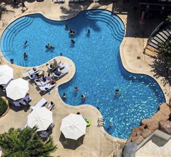 En vy från ovan av en pool med en ovanlig form, omgiven av solstolar och parasoller, med en stenlagd gångväg som leder fram till den.