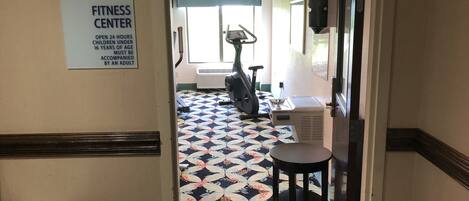 Een fitnessruimte met een patroon vloer, trainingsapparatuur en een bord waarop staat dat deze 24 uur per dag geopend is voor kinderen onder de 16 jaar onder begeleiding van een volwassene.