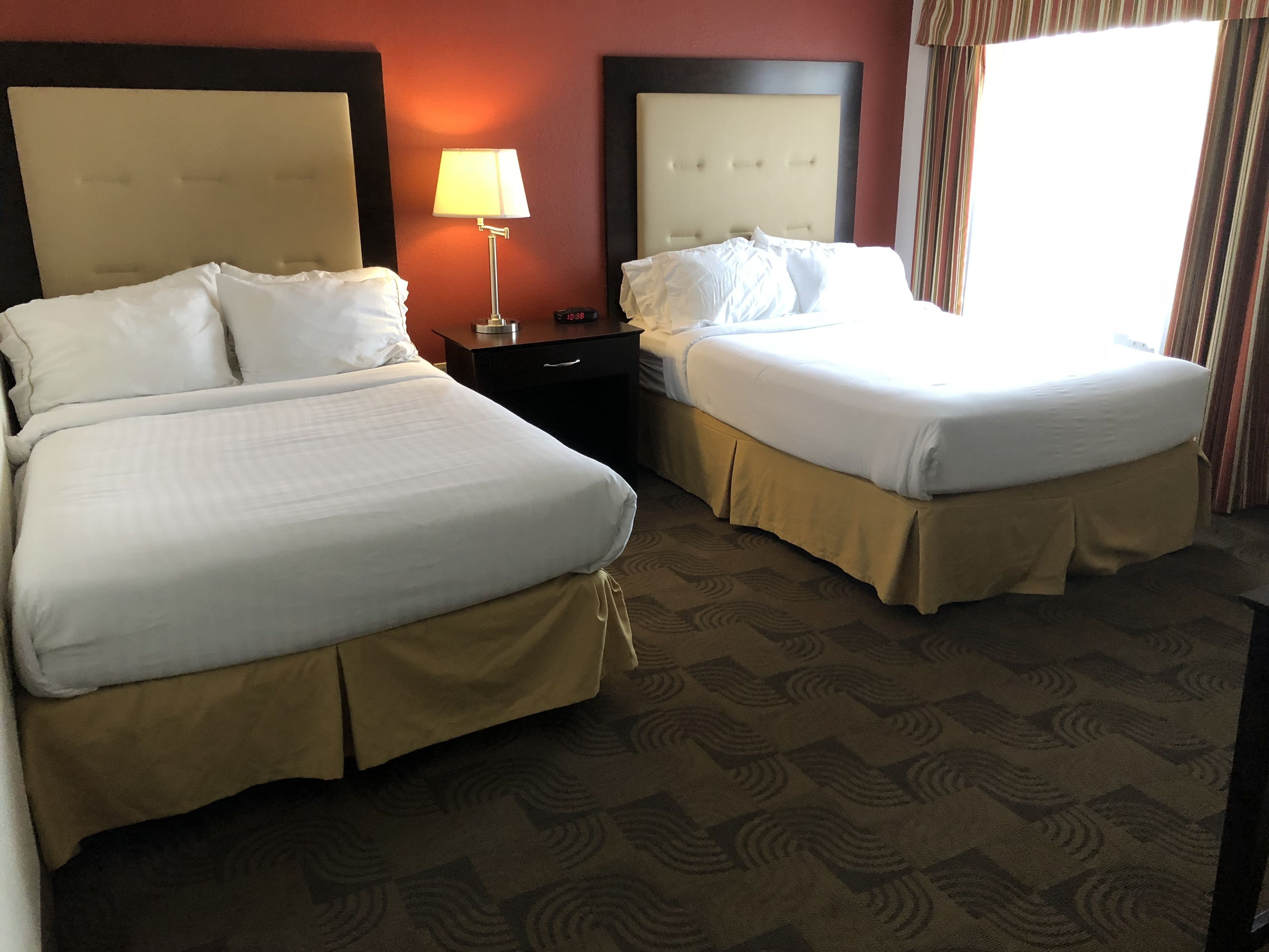 A hotel room with two beds, a nightstand, a lamp, and a window with curtains.