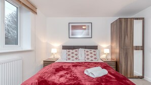 Een net opgemaakt bed met een rode deken en een opgerolde handdoek, twee bedlampen, een ingelijste foto aan de muur en een garderobe met spiegel.