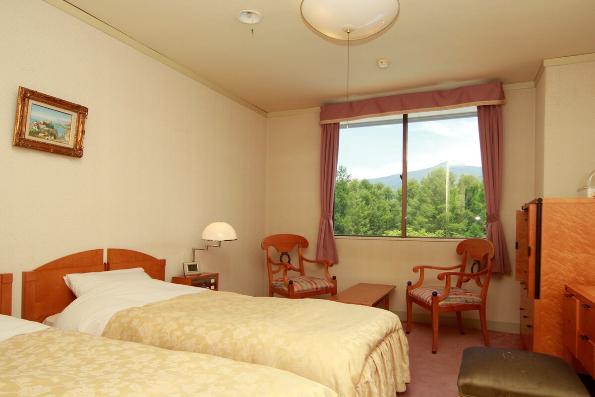 A hotel room with two beds, a desk, a chair, a lamp, and a window with a view of trees.