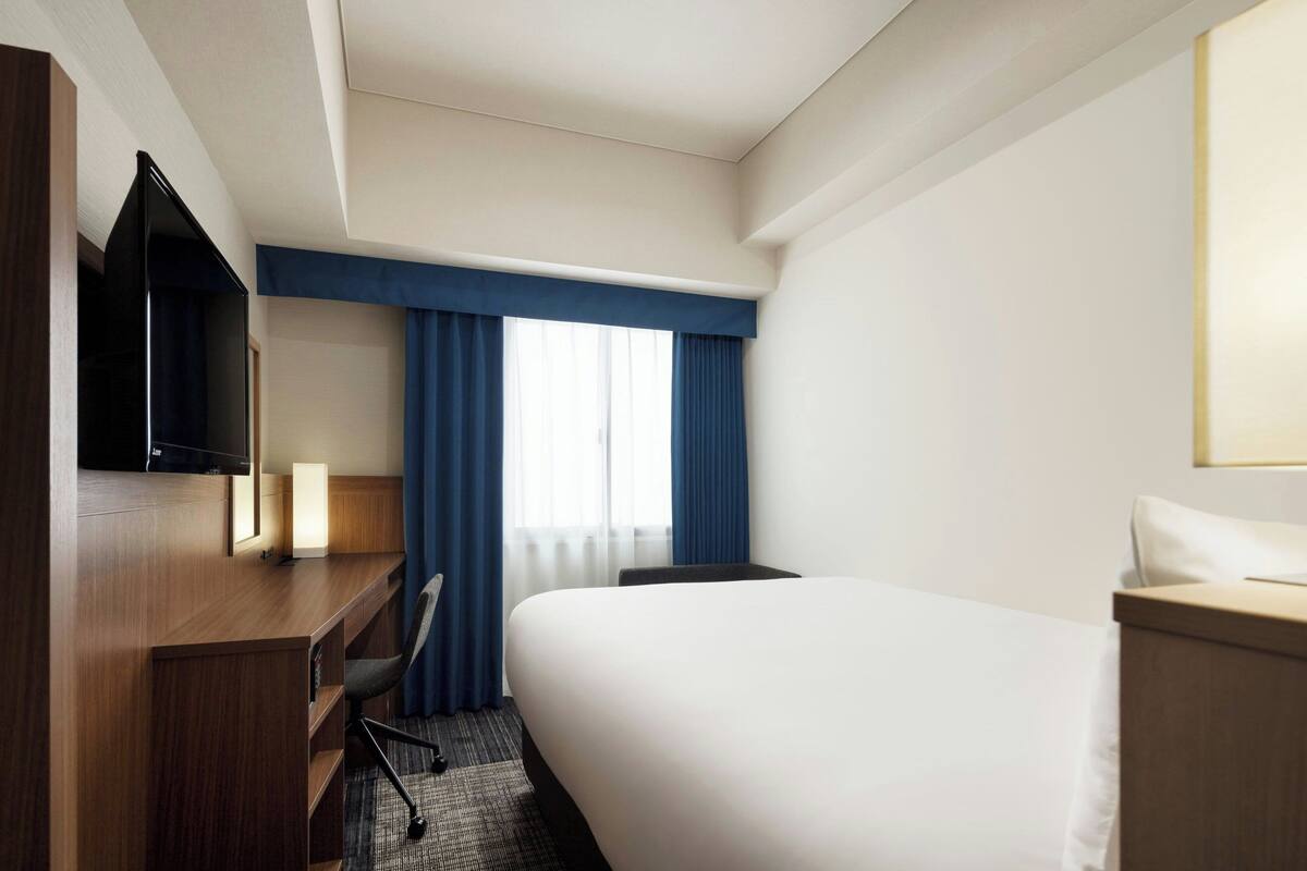 A hotel room with a bed, a desk with a chair, a television, and a window with curtains.