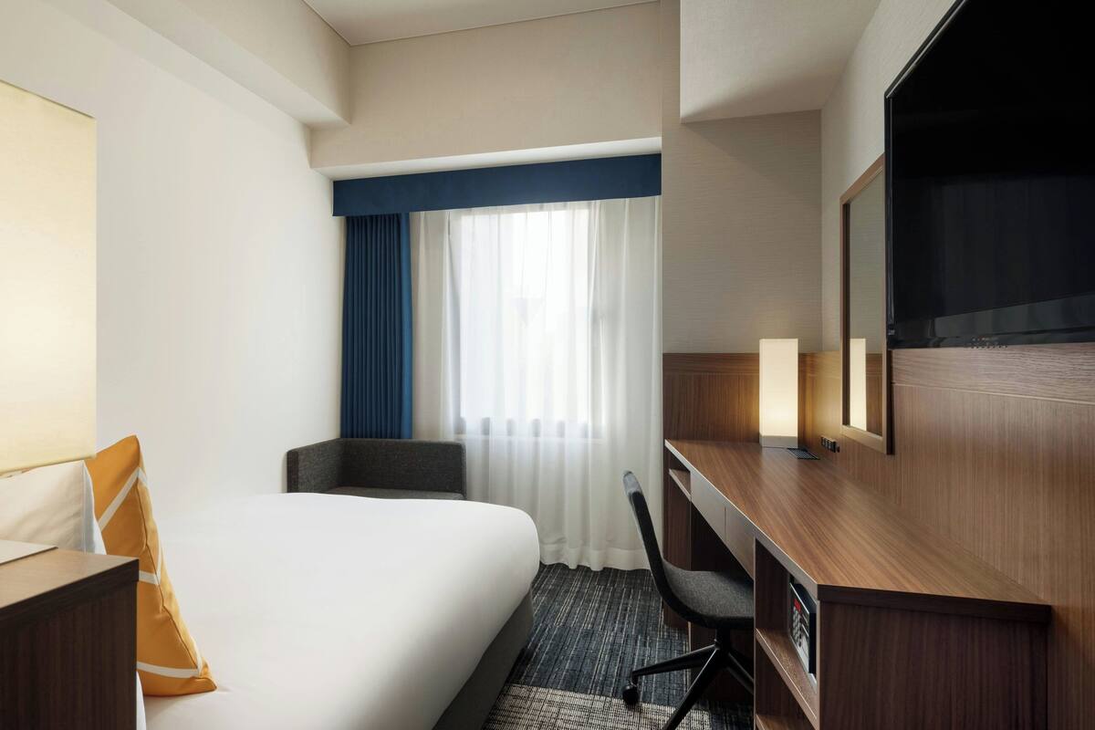 A hotel room with a bed, a desk, a chair, a TV, and a window with curtains.