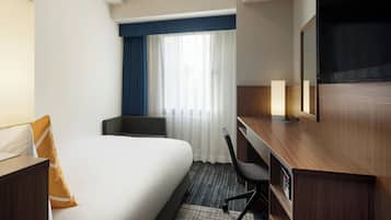 Quarto de hotel com cama, escrivaninha, cadeira, televisão e janela com cortinas.