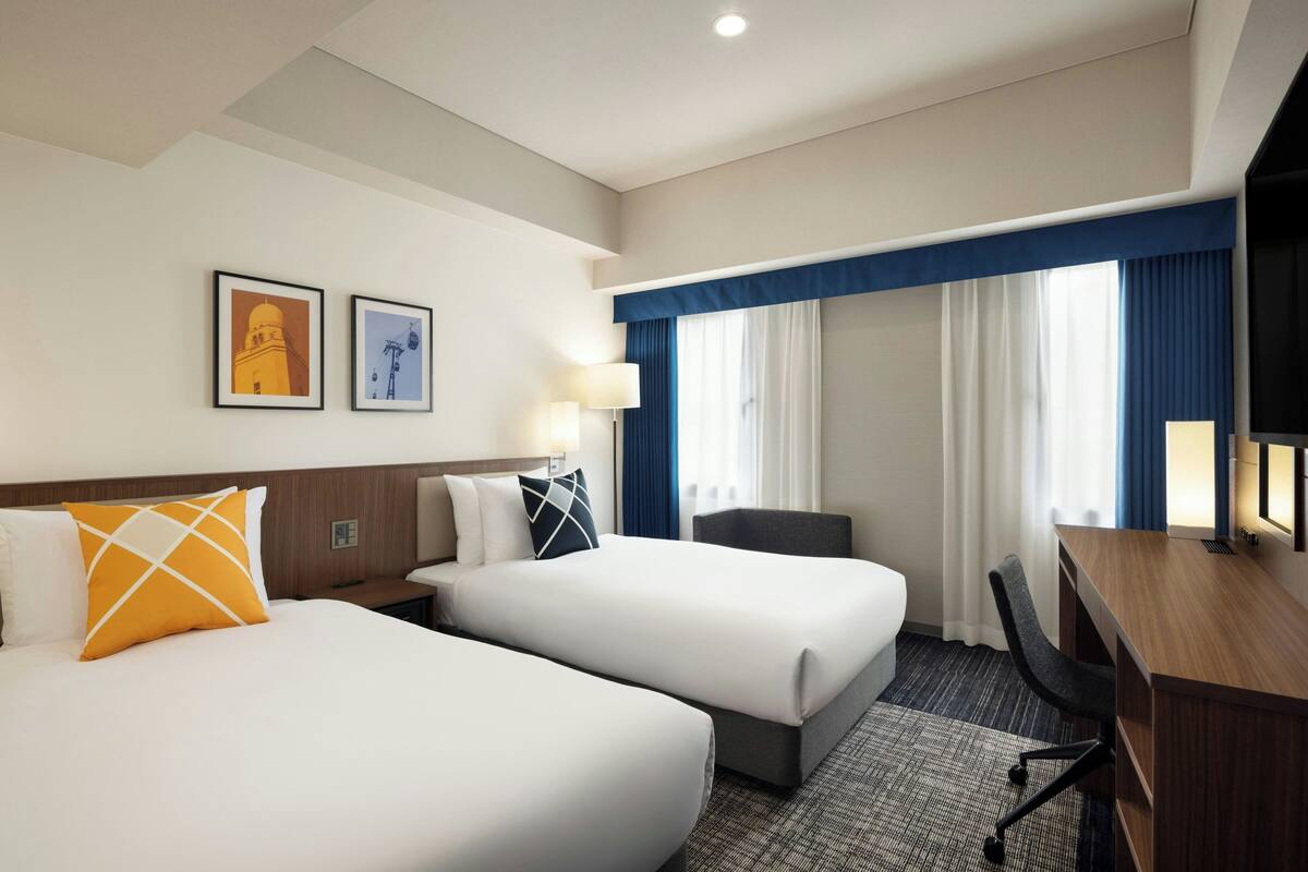 A hotel room with two beds, a desk, and a chair. There are two framed pictures on the wall.