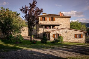 Una casa de piedra con balcón, rodeada de vegetación y un camino de grava.