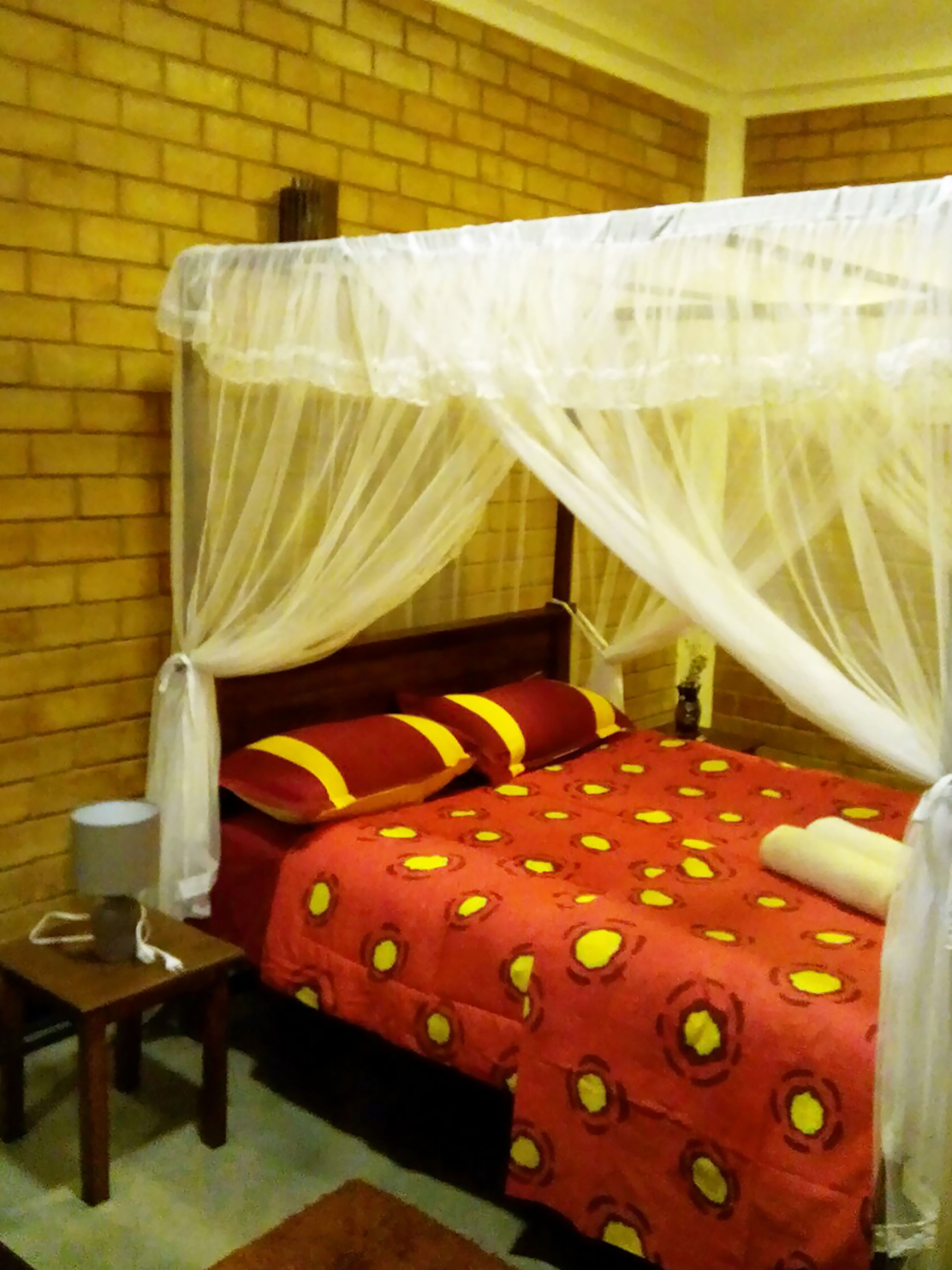 A room with a brick wall, a bed with a red bedspread and yellow pillows, a nightstand with a lamp, and a white canopy over the bed.