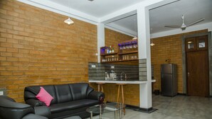 A modern living room with a brick wall, a black leather sofa, a glass coffee table, and a kitchen area with a refrigerator and shelves. - Brown House (Unawatuna)