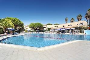 Ein Schwimmbecken mit klarem Wasser, umgeben von Liegestühlen und Sonnenschirmen, mit Palmen und Gebäuden im Hintergrund.