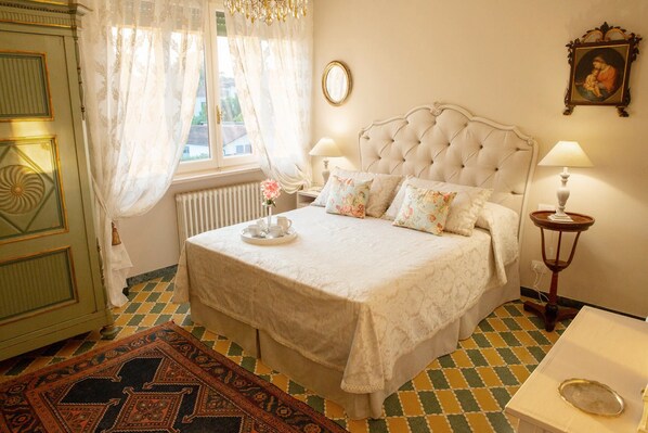 A bedroom with a large bed, two bedside tables, a window with curtains, a radiator, and a patterned rug.