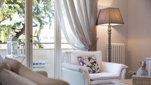 A living room with a white armchair, a table with a lamp, and a view of the outdoors through a window with curtains.