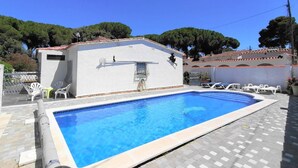 Una piscina rectangular con borde de baldosas, rodeada de sillas de descanso y un pequeño edificio con techo de tejas.