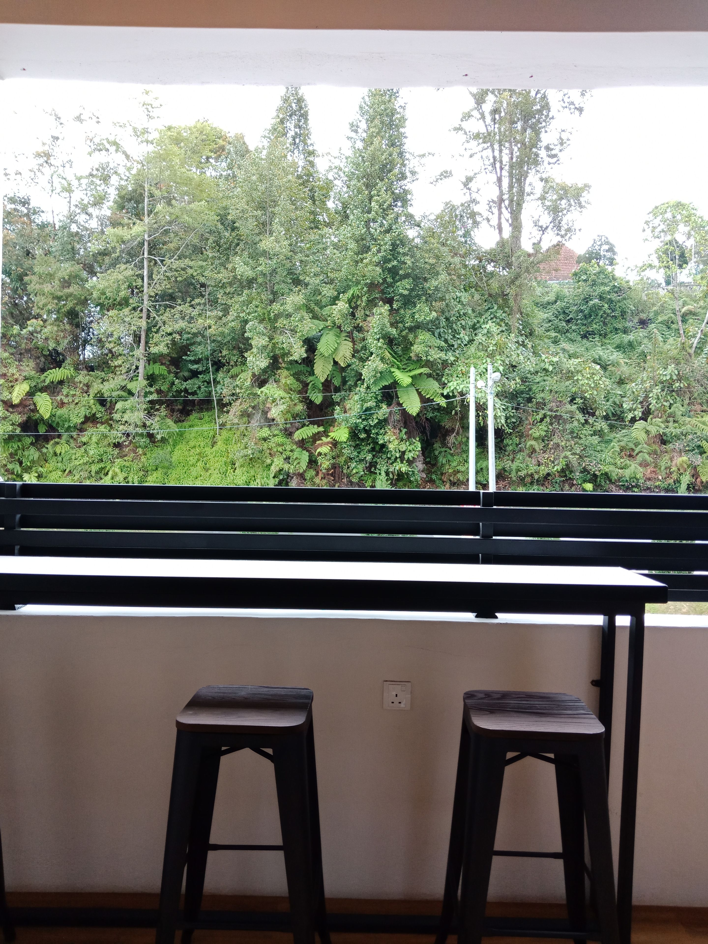 A balcony with a view of dense greenery and trees. There are two black stools and a white table.