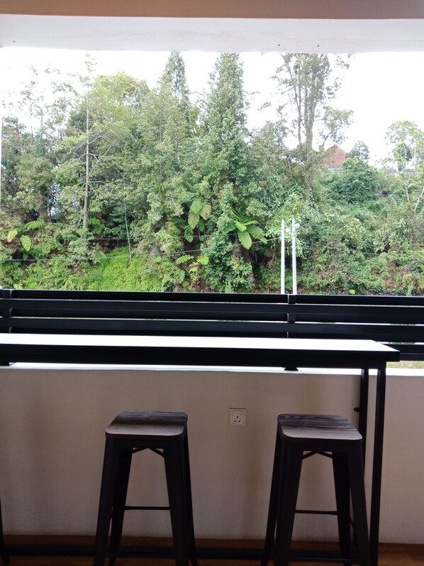 A balcony with a view of dense greenery and trees. There are two black stools and a white table.
