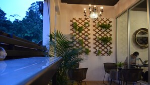 Un rincón interior acogedor con un banco de madera, dos sillas de mimbre y una mesa. Una pared de celosía de madera con plantas en macetas decora el espacio.