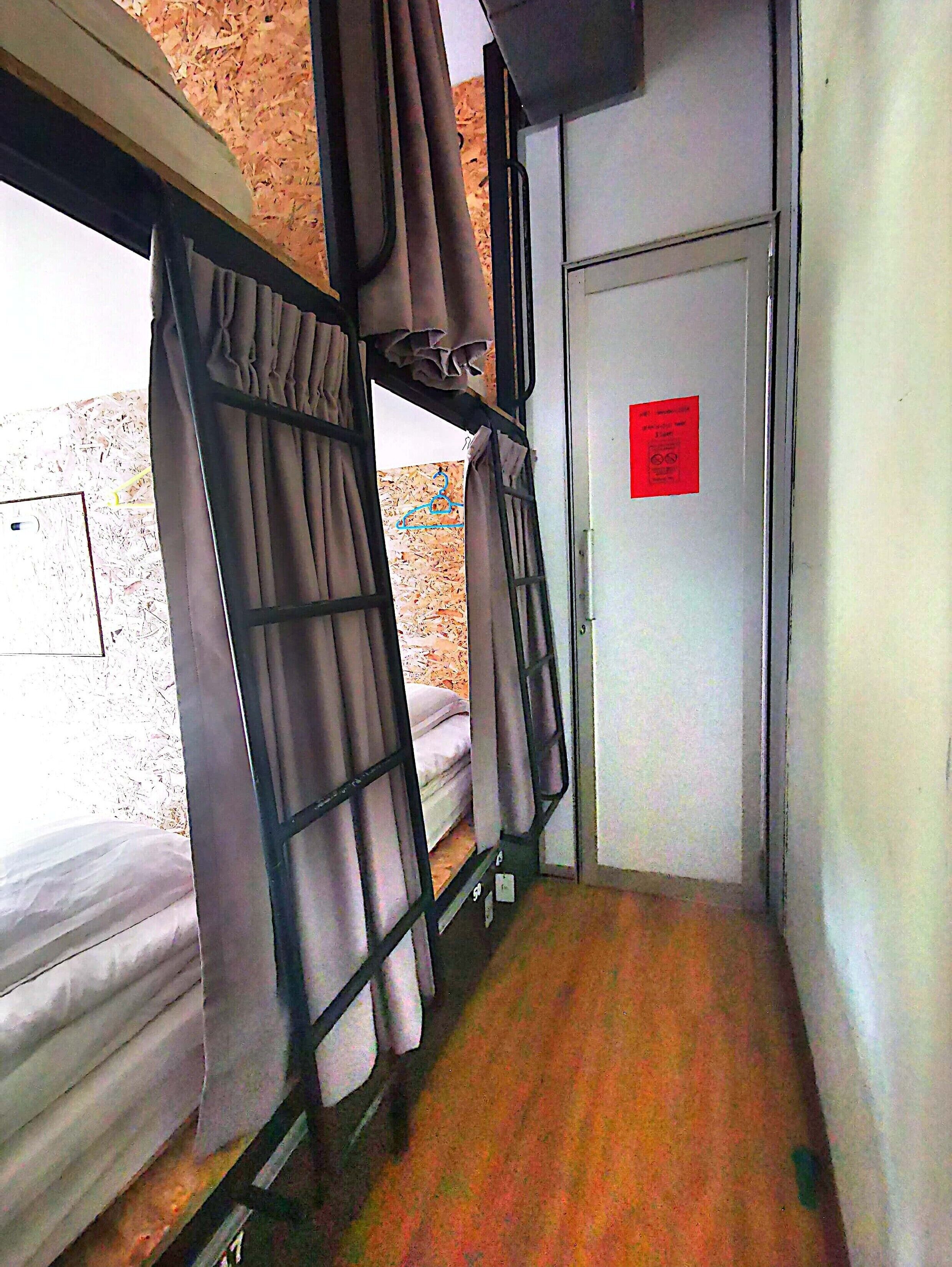 Un estrecho pasillo con literas a ambos lados, una puerta con un letrero rojo y una ventana con cortinas.