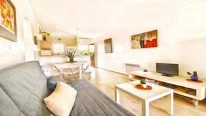 Ein modernes Wohnzimmer mit einem grauen Sofa, einem kleinen Tisch und einem Fernseher. Im Hintergrund befindet sich eine Küchenzeile mit einem Esstisch und Stühlen.