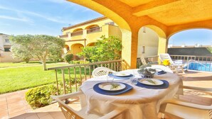 Eine Terrasse mit Tisch für zwei Personen, mit Blick auf einen Pool und ein gelbes Gebäude mit Bögen.