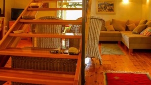 Een houten trap leidt naar een hoger gelegen verdieping met een balkon, een woonkamer met een bank en een televisie, en een rode vloerkleed.
