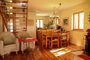 A cozy living room with wicker furniture, a dining table, and a kitchen area with wooden cabinets and a chandelier.