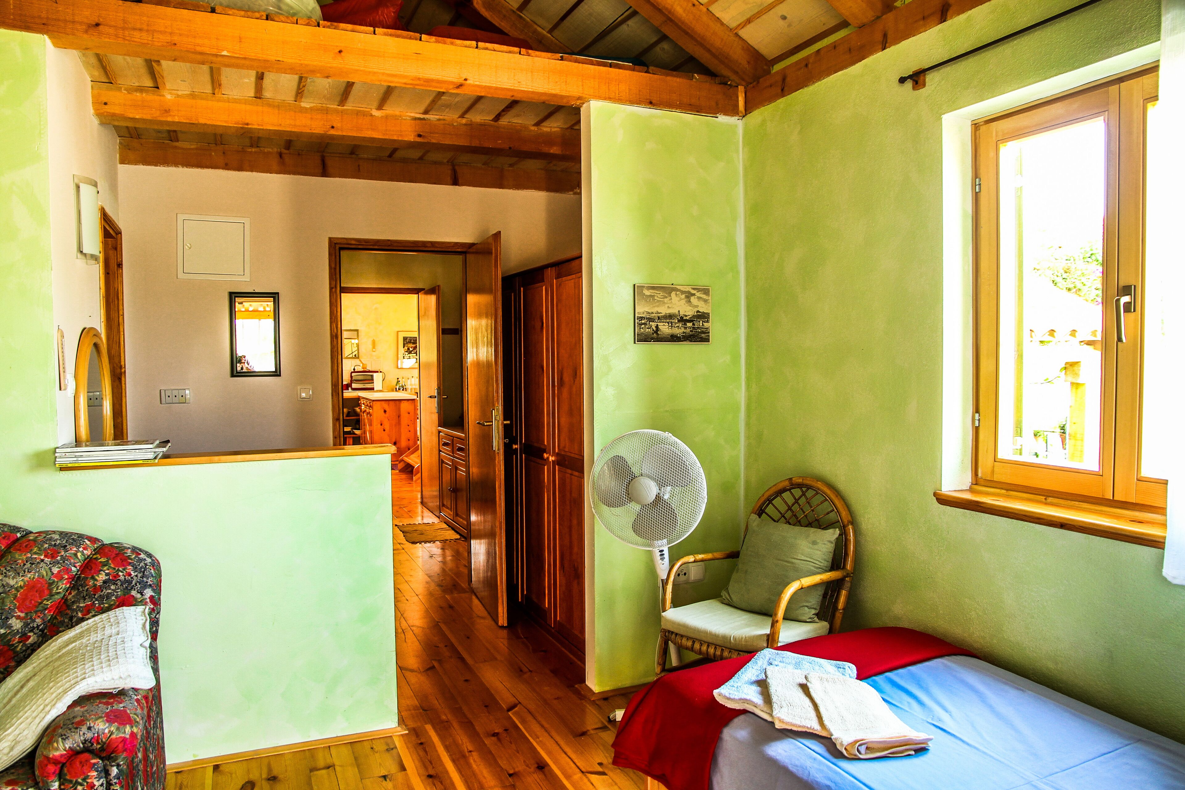 Una habitación acogedora con piso de madera, una cama con manta roja, una silla, un ventilador de pie y una ventana con vistas al exterior.