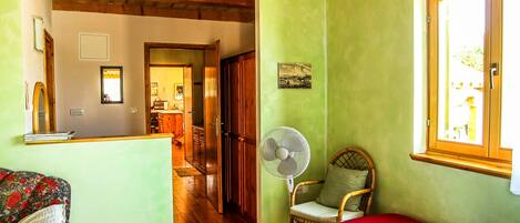 Una habitación acogedora con piso de madera, una cama con manta roja, una silla, un ventilador de pie y una ventana con vistas al exterior.