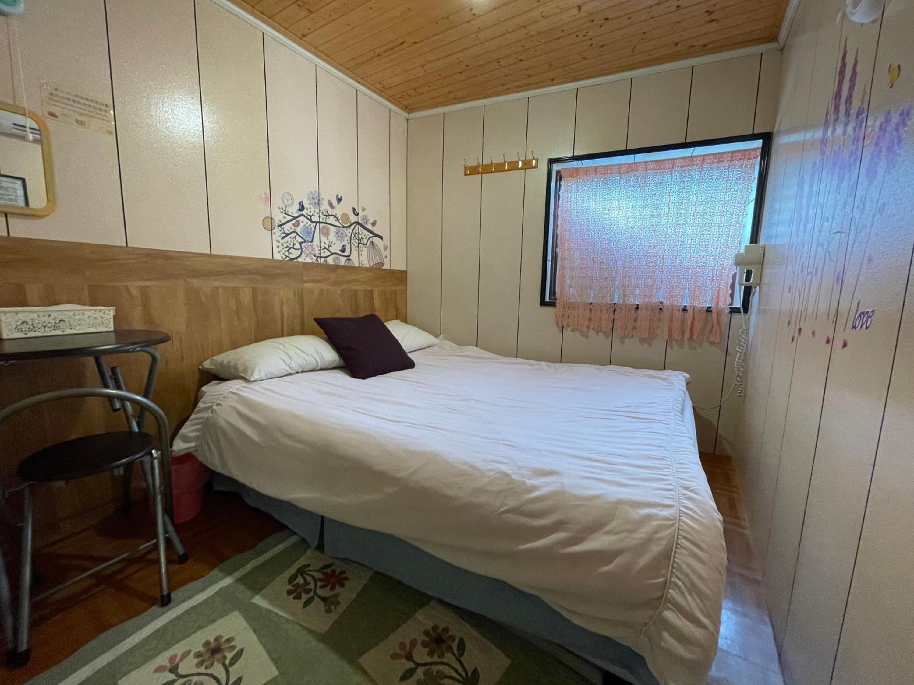 A single bed with a headboard, a small table with a chair, a window with lace curtains, and a wall with a decorative piece and a sign.