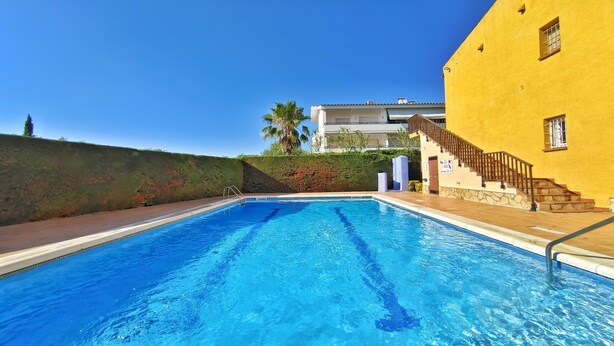 A swimming pool with a clear blue water, surrounded by a wooden deck. There is a staircase leading to a yellow building with a balcony. - VACAY L'Escala - Apartamento Tropicana (L'ESCALA)