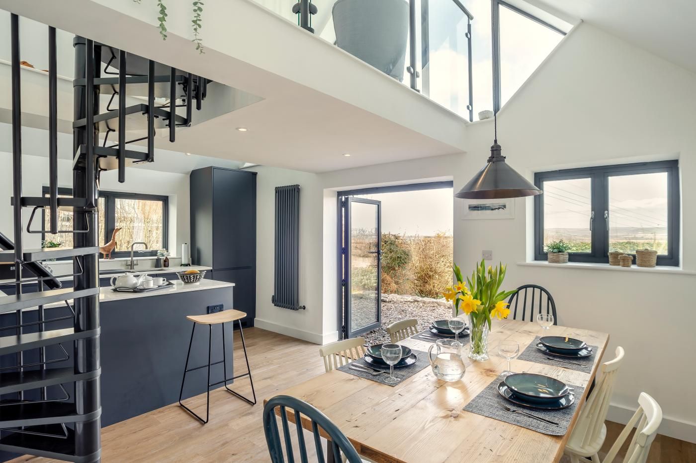 A modern kitchen with a central island, a dining area with a wooden table, and a view of the outdoors through large windows.