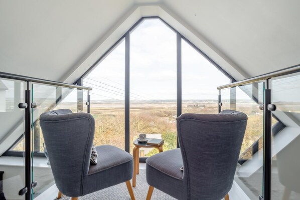 Una habitación moderna con un tragaluz triangular grande, dos sillones tapizados en gris y una mesita con una taza y un libro.