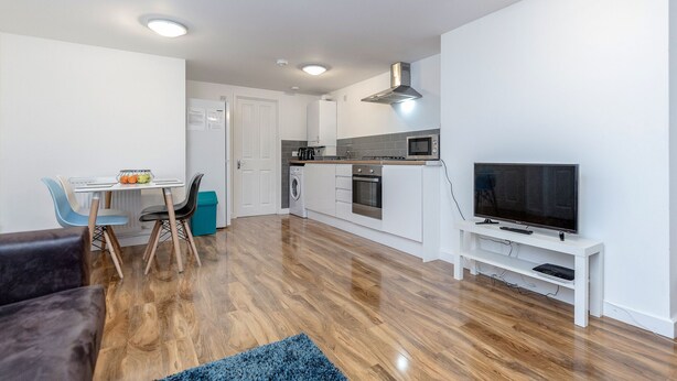 A modern kitchen with white cabinets, a microwave, and a stove. There is a dining area with a table and chairs, and a TV on a stand. - Royal Apartments RA2 (Greater London)