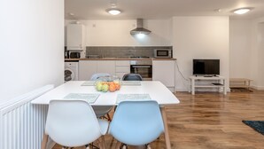A modern kitchen with white cabinets, a microwave, and a stove. There is a dining table with chairs and a bowl of fruit. A TV is on a stand in the corner. - Royal Apartments RA2 (Greater London)