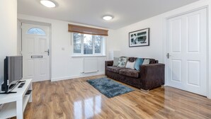 A modern living room with hardwood flooring, a brown sofa with cushions, a television on a white stand, a window with blinds, and a framed picture on the wall. - Royal Apartments RA2 (Greater London)