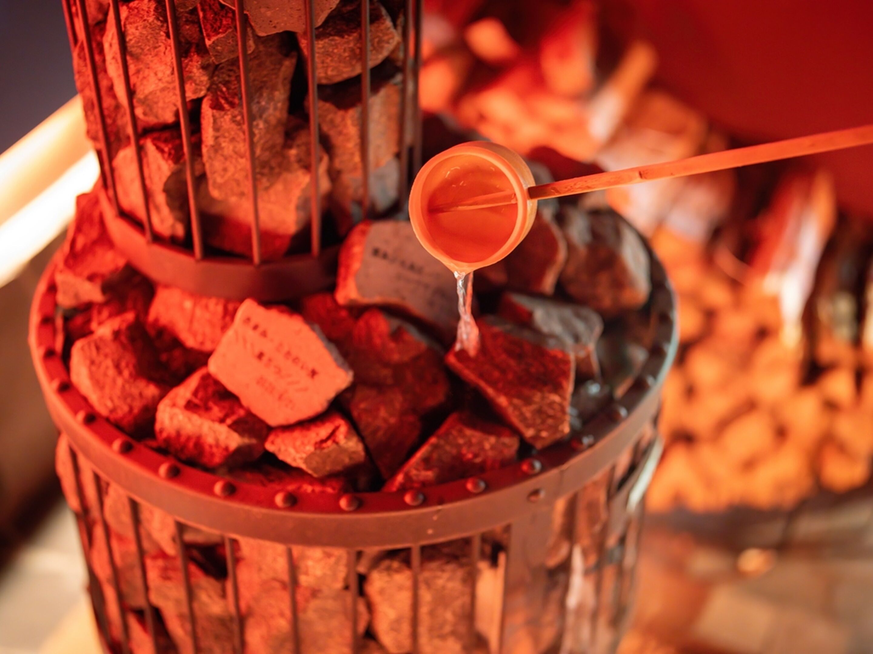 A close-up of a charcoal grill with a metal rack holding charcoal pieces, and a metal spoon with a liquid being poured over the charcoal.