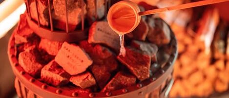 A close-up of a charcoal grill with a metal rack holding charcoal pieces, and a metal spoon with a liquid being poured over the charcoal.