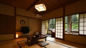 Ein traditioneller japanischer Raum mit Tatami-Boden, einem niedrigen Tisch, einem Fernseher und großen Fenstern mit Blick ins Grüne.