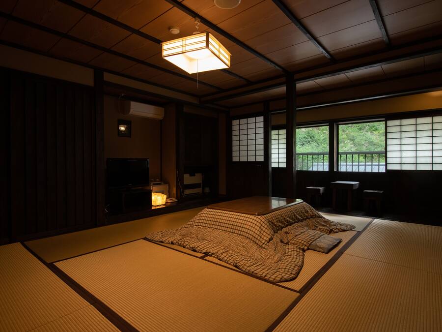 Une pièce traditionnelle avec un sol recouvert de tatamis, un plafond bas, des portes coulissantes et une vue sur la verdure à l’extérieur.