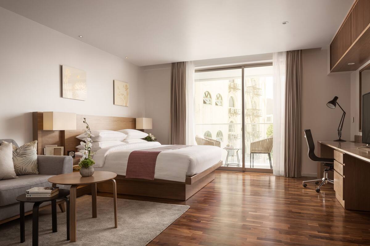 A modern hotel room with a bed, a desk, a chair, a sofa, and a balcony with a view of the city.