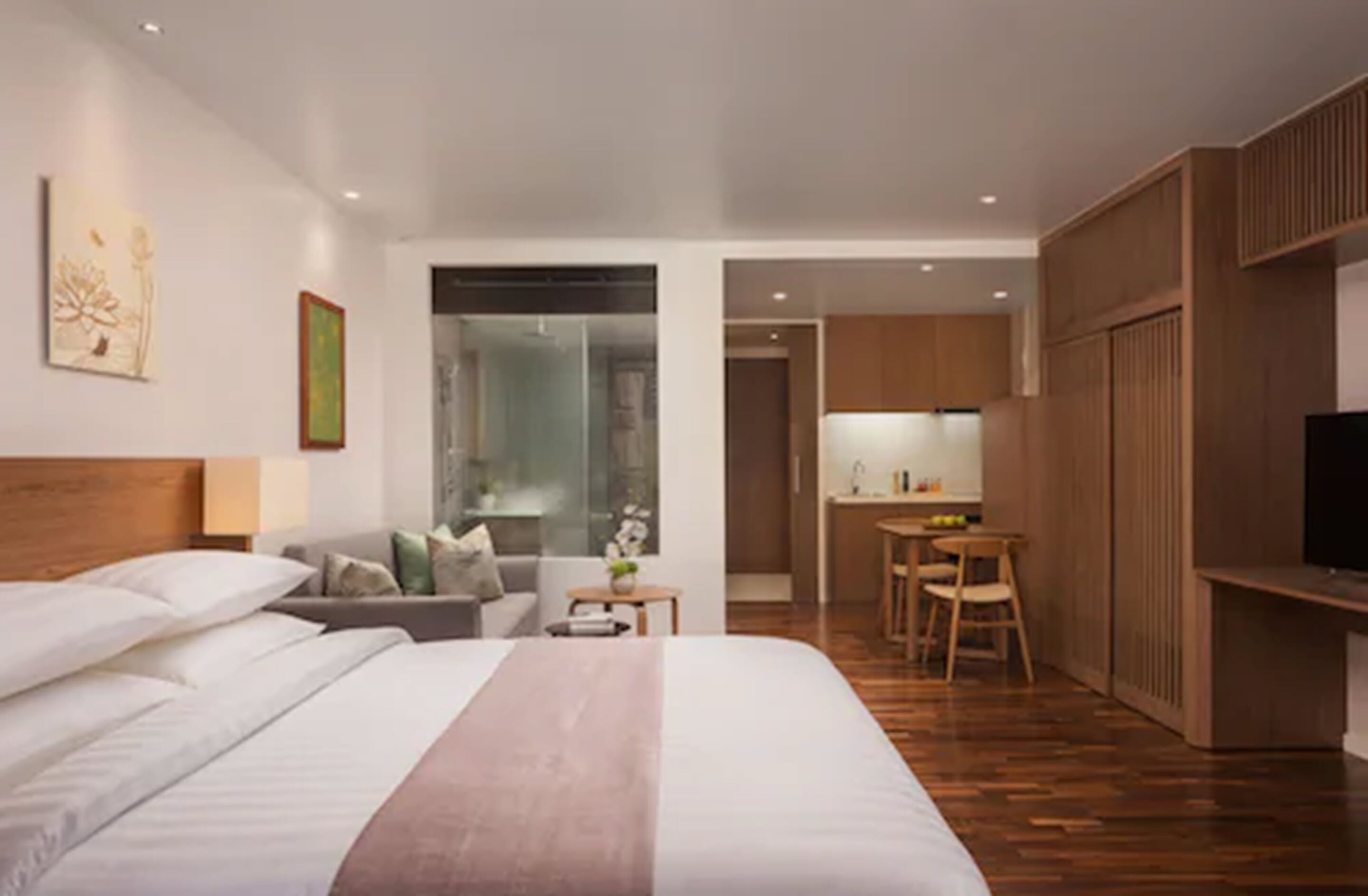 A modern hotel room with a large bed, a sofa, a small table, a TV, and a kitchenette.