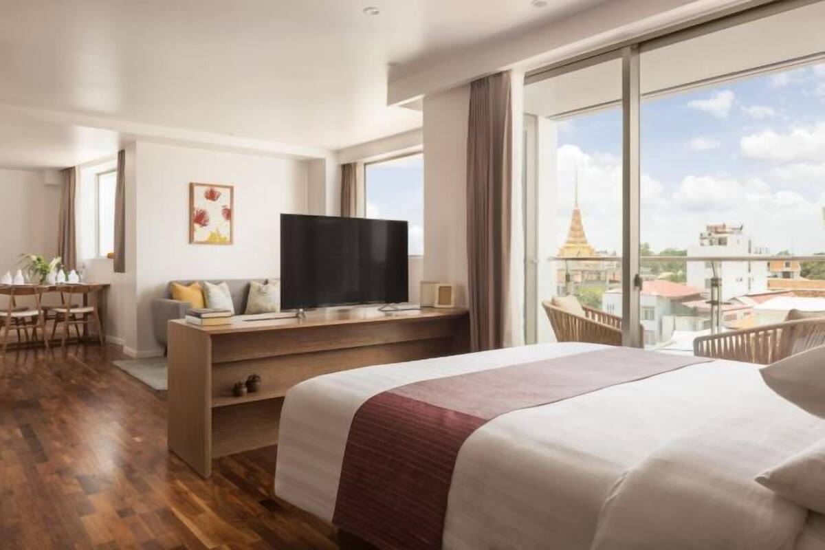 A hotel room with a bed, a TV on a wooden stand, a desk with a chair, and a large window with a view of buildings and a temple.
