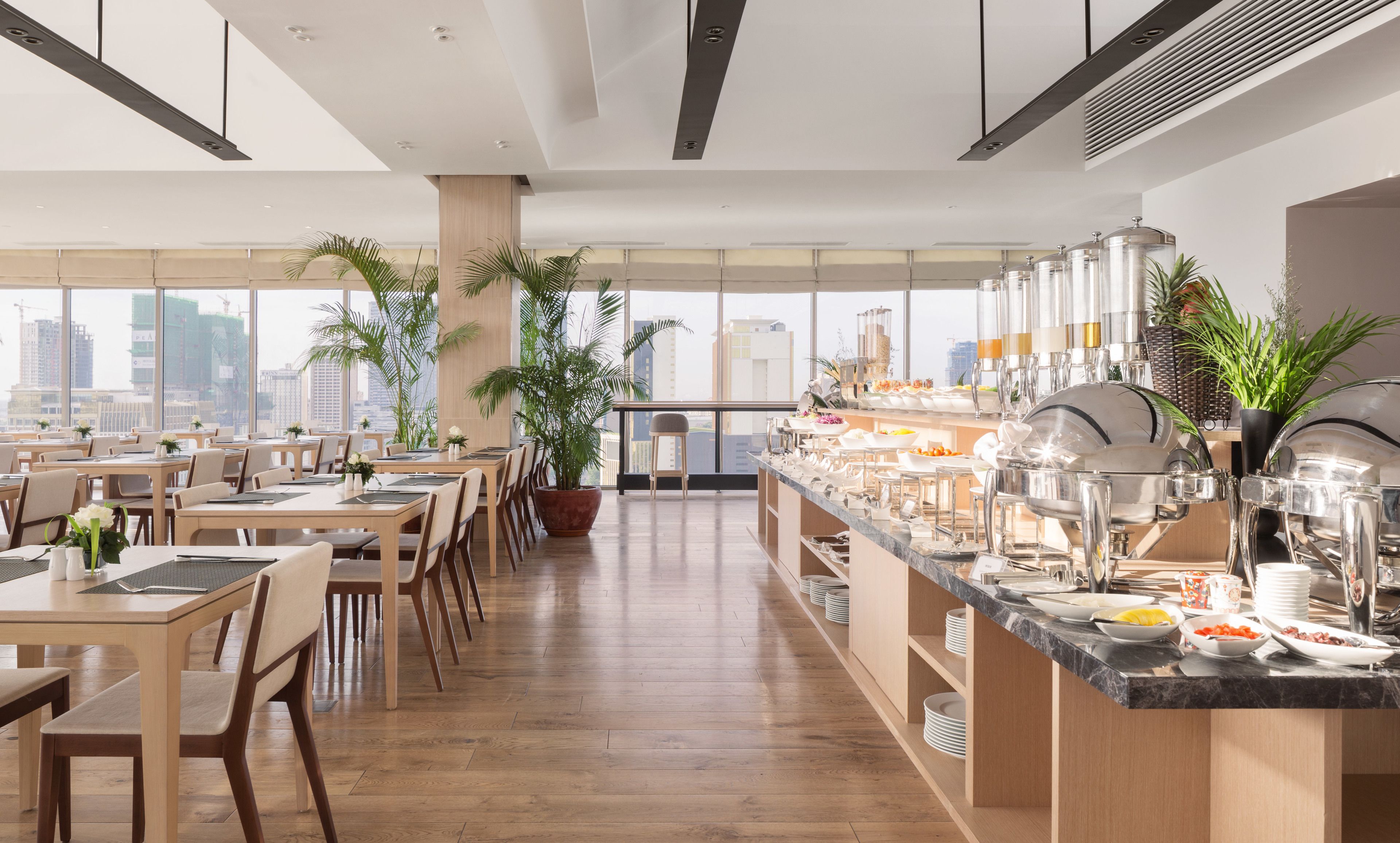 A modern dining area with wooden tables and chairs, a buffet counter with various food items, and large windows offering a city view.