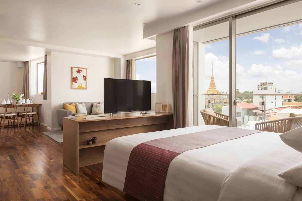A hotel room with a large bed, a desk with a TV, a balcony with a view of a temple and buildings.